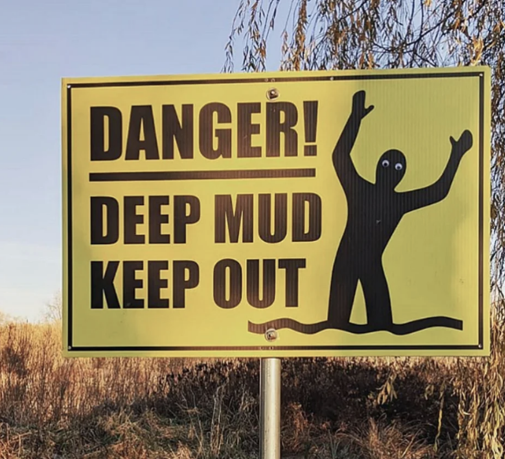 Sign with a figure warning, "Danger! Deep Mud Keep Out" against a rural background with tall grass and a tree