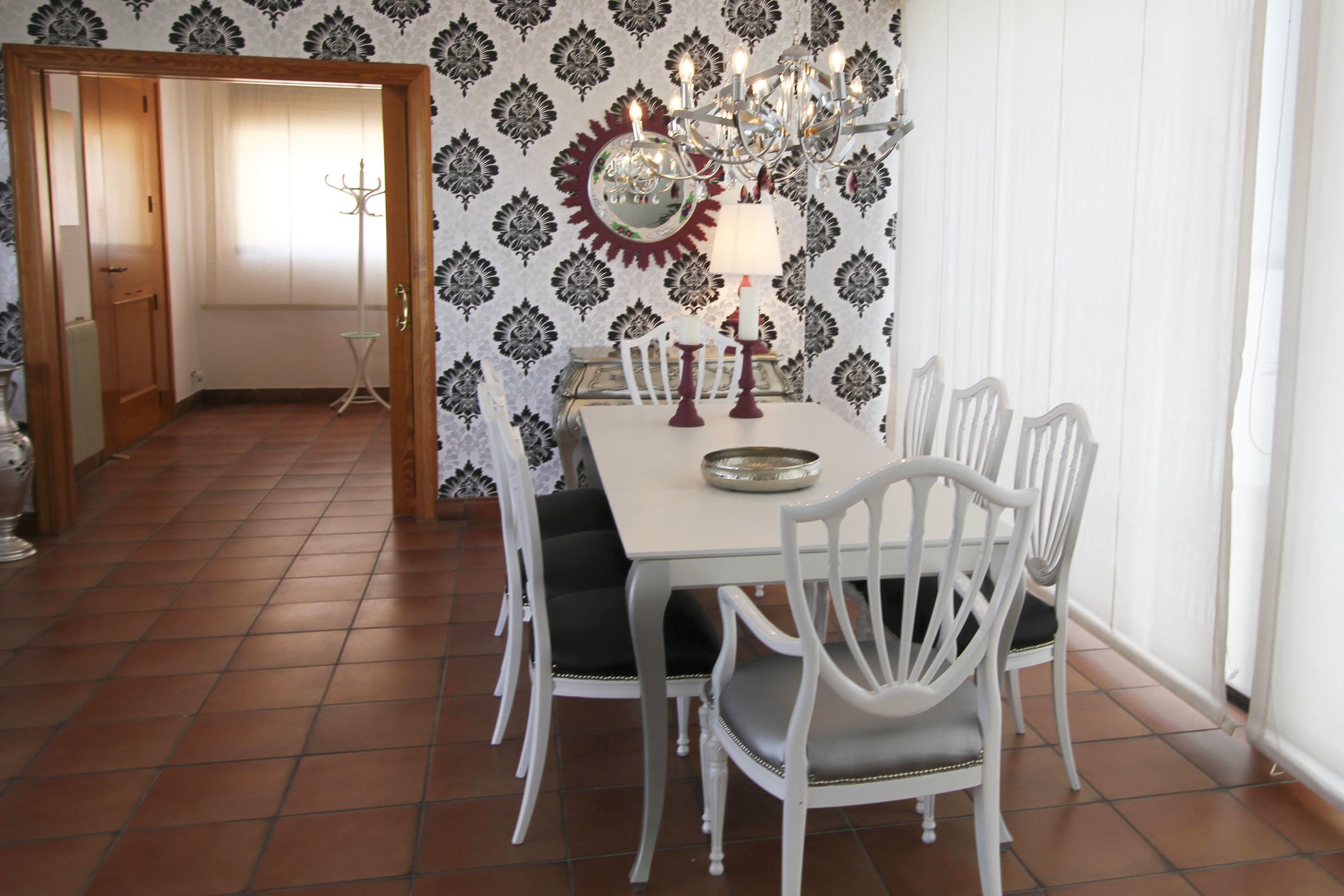 A stylish dining room with a white table, six white chairs, a decorative wall, and a chandelier. The background shows an adjacent room with tiled flooring
