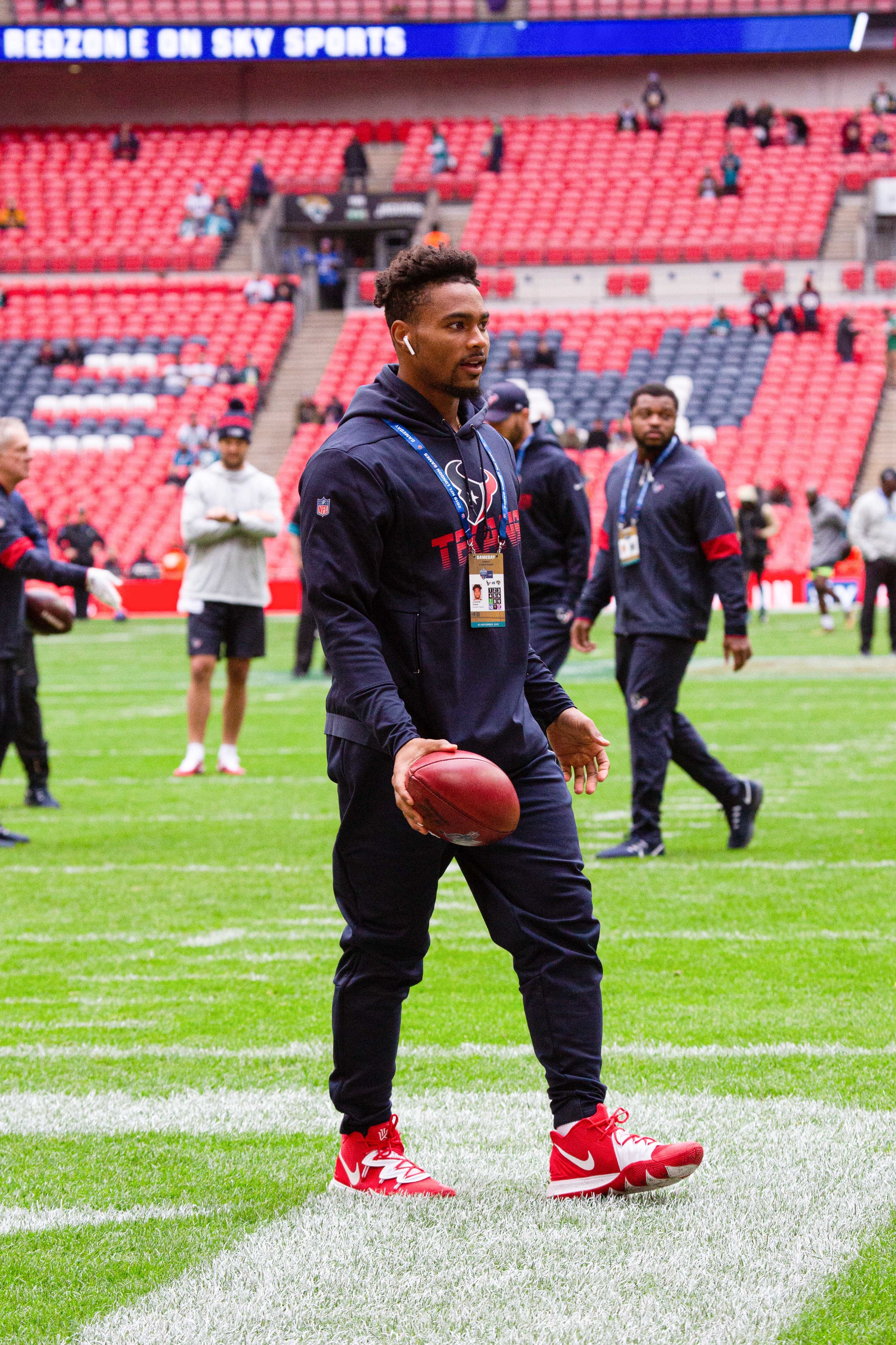 A football player in warm-up gear holding a football on a field with other team members and staff in the background at a stadium