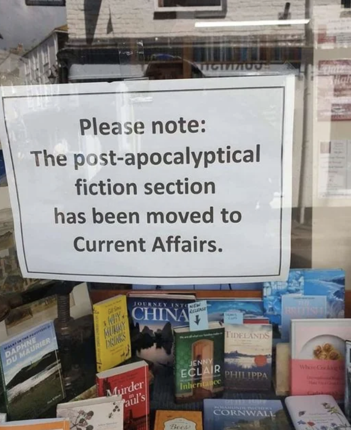 A bookstore sign reads: "Please note: The post-apocalyptical fiction section has been moved to Current Affairs." Books are displayed below the sign