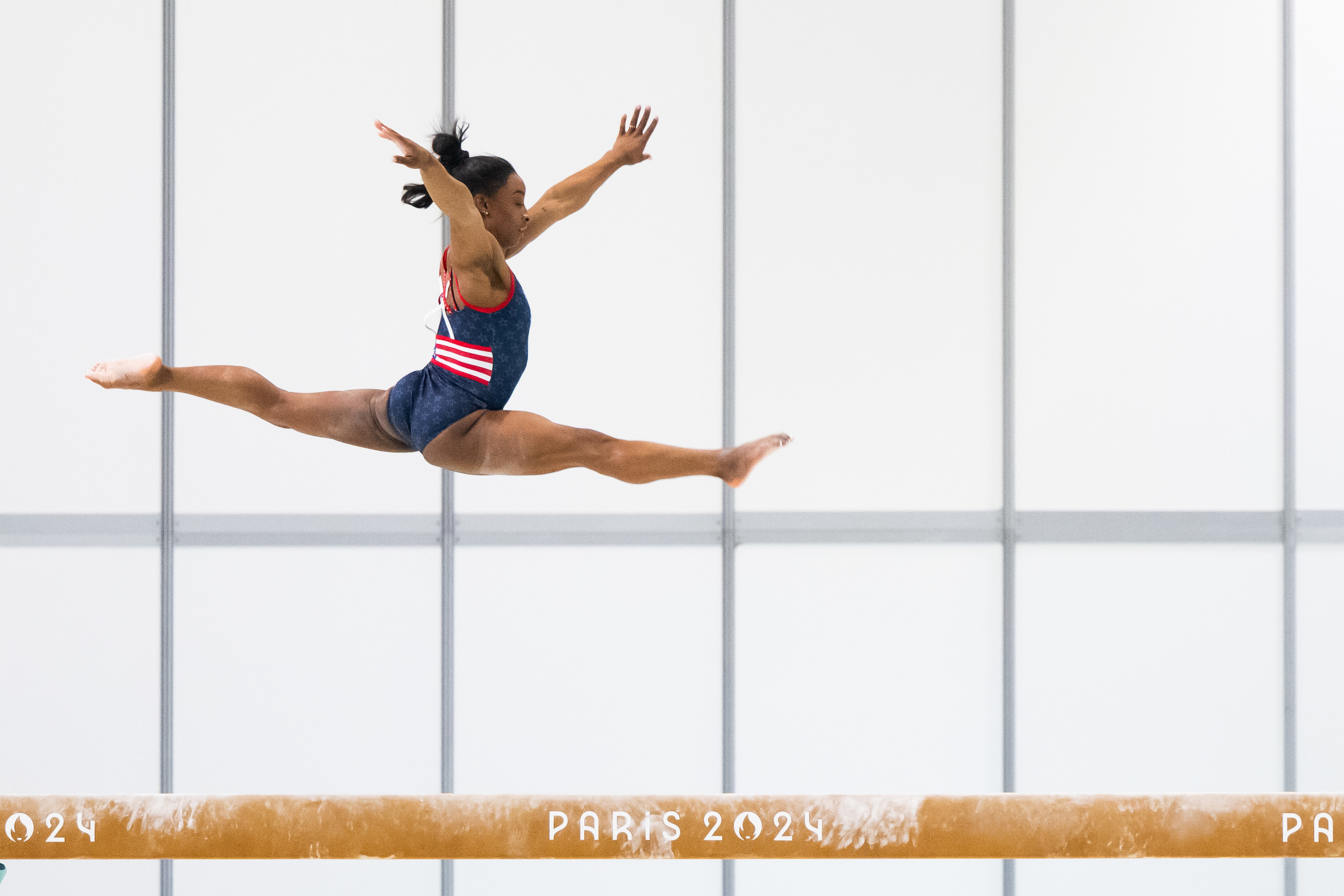 Simone Biles performs a split leap in the air above a balance beam labeled &quot;Paris 2024&quot; during a gymnastics competition