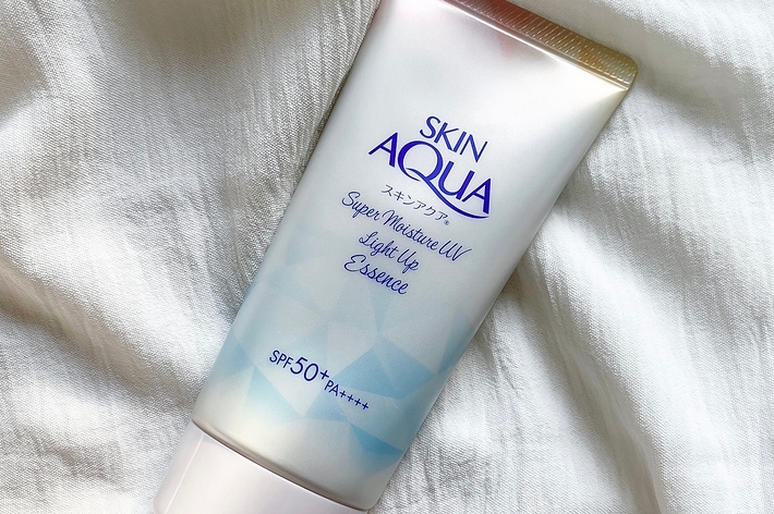 SKIN AQUAのスーパー保湿UVライトアップエッセンス、SPF50+ PA++++入り、白い生地の上に置かれている。