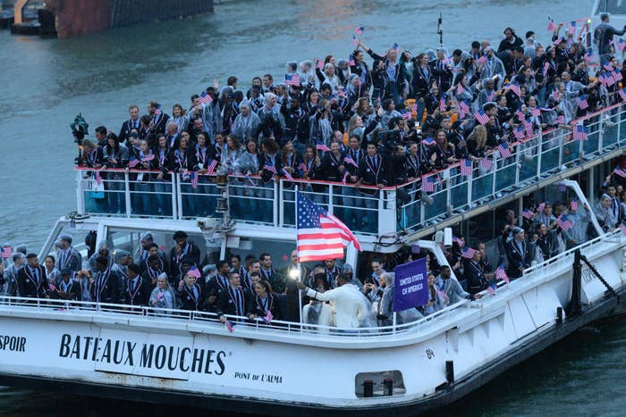 A boat crowded with people in formal clothing holds American and French flags while sailing. People are celebrating on the Bateaux Mouches in Paris
