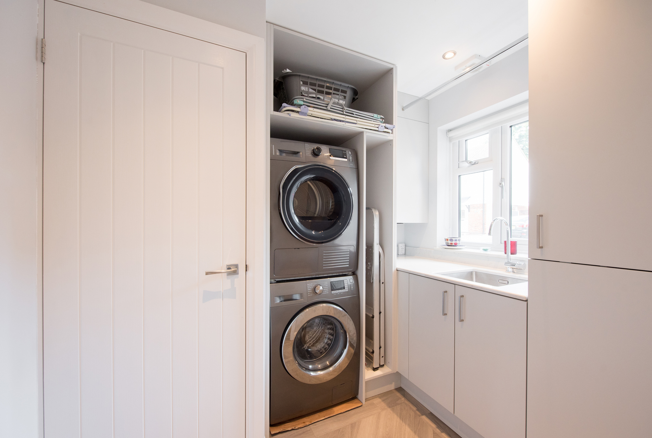 A modern laundry room features a stacked washer and dryer, surrounding cabinets, a folding area with a laundry basket, and a sink next to a window