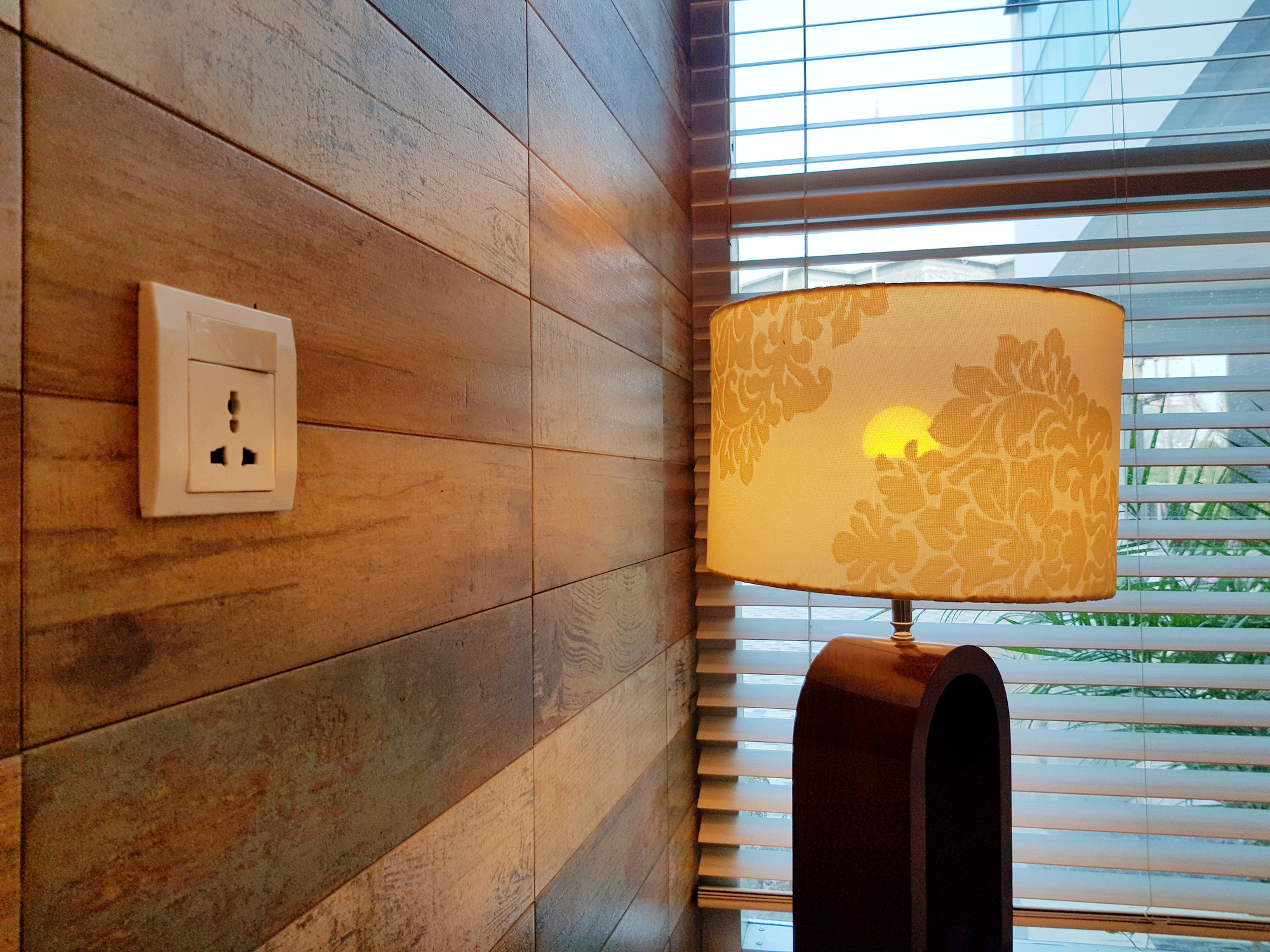 A wooden wall with an electrical outlet next to a lamp with a floral-patterned lampshade, placed by a window covered with blinds