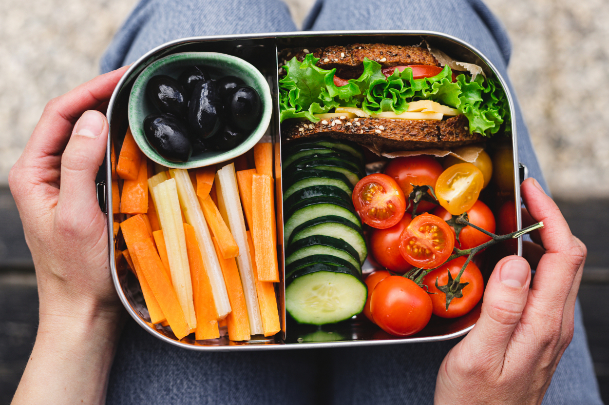 Someone holding a lunchbox that has carrot sticks, black olives, a sandwich with lettuce and tomato, cucumber slices, and cherry tomatoes in it