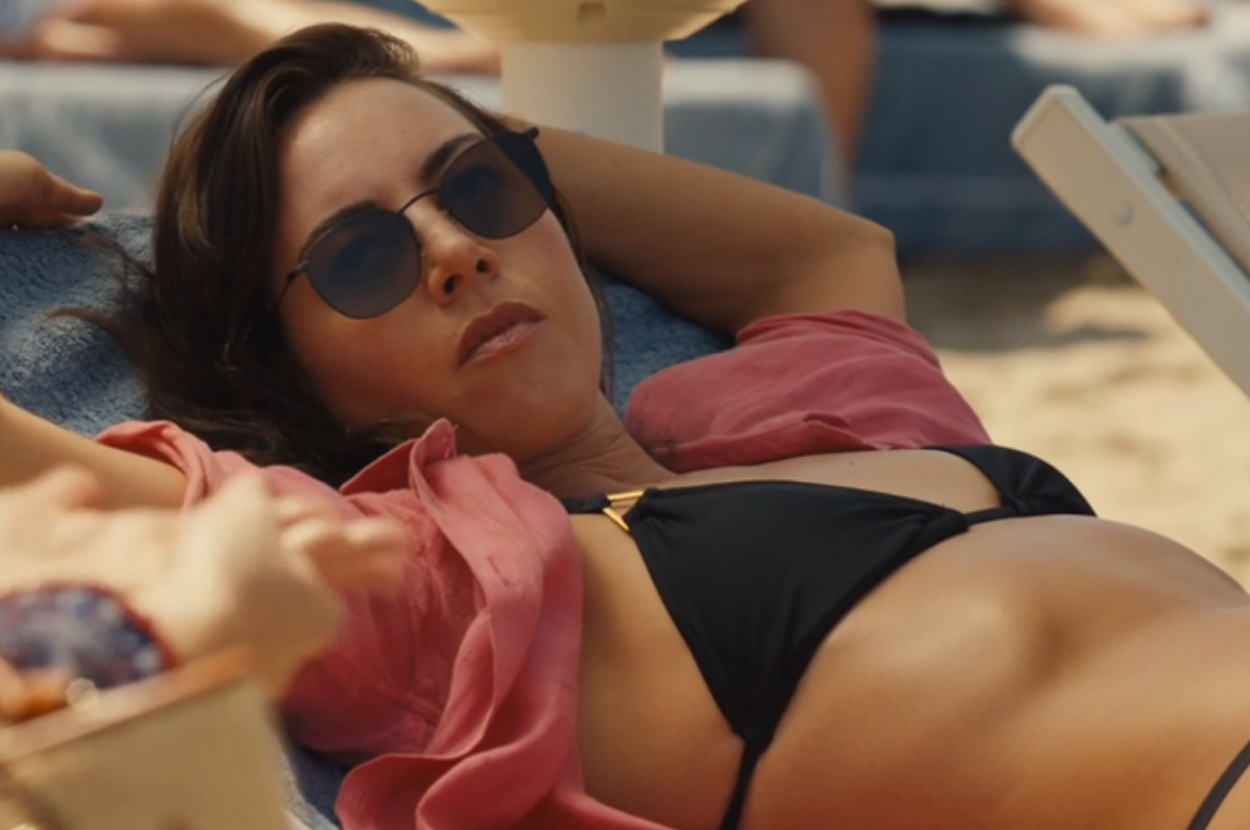 Aubrey Plaza is lounging on a sunbed at the beach, wearing sunglasses, a dark bikini, and an open shirt as Harper on The White Lotus