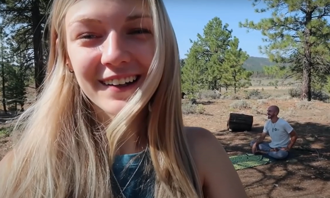 Gabby Petito smiles at the camera while a man sits on a mat, outdoors in a wooded area.