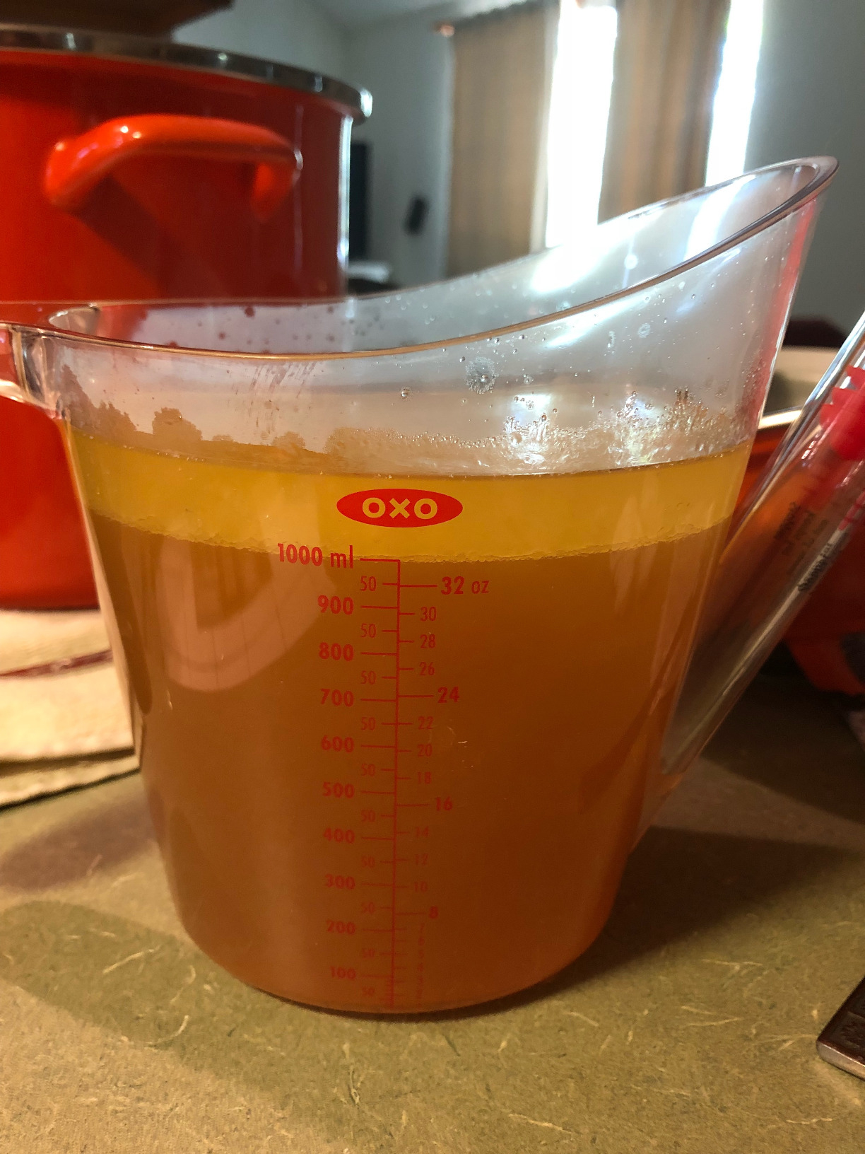 A clear measuring cup filled with broth on a kitchen countertop, with a pot and curtains in the background