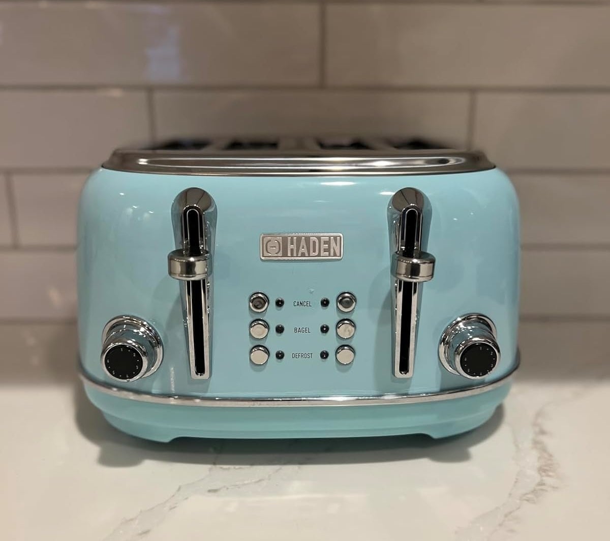 A Haden brand toaster with four slots is placed on a kitchen counter with a tiled backsplash. The toaster has settings for cancel, bagel, and defrost
