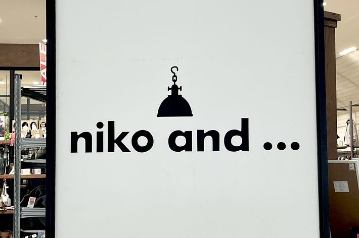 Storefront sign displaying &ldquo;niko and &hellip; &quot; in black text with a small hanging lamp graphic above the text. Shelves with various products are visible in the background