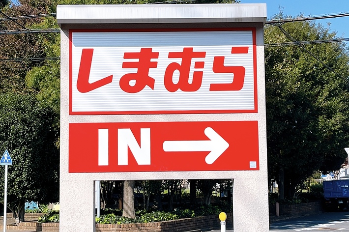 A bilingual sign with Japanese characters and an arrow pointing right. The sign reads &ldquo;Shimamura&rdquo; and &ldquo;IN&rdquo; indicating direction. Trees and road in background