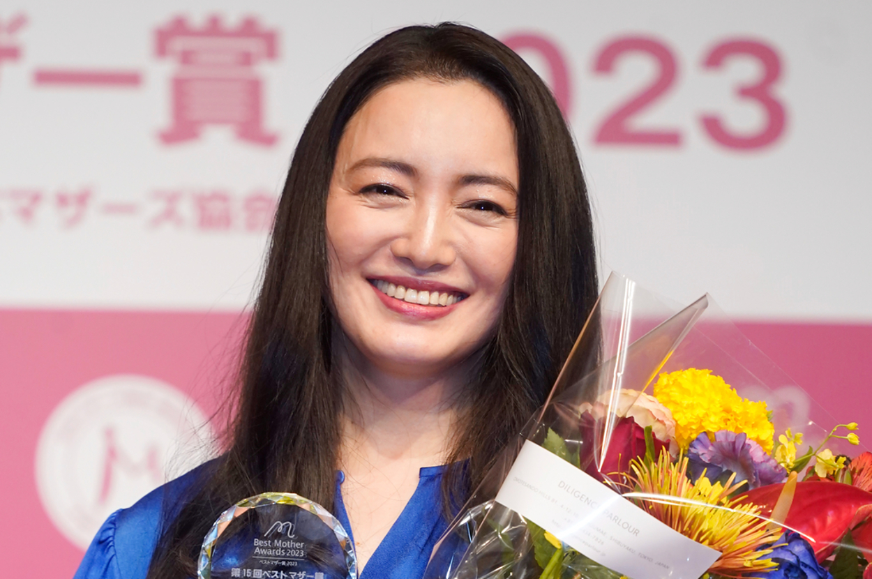 女優の石原さとみが、青いドレスを着てトロフィーと花束を手に笑顔を見せているシーン。