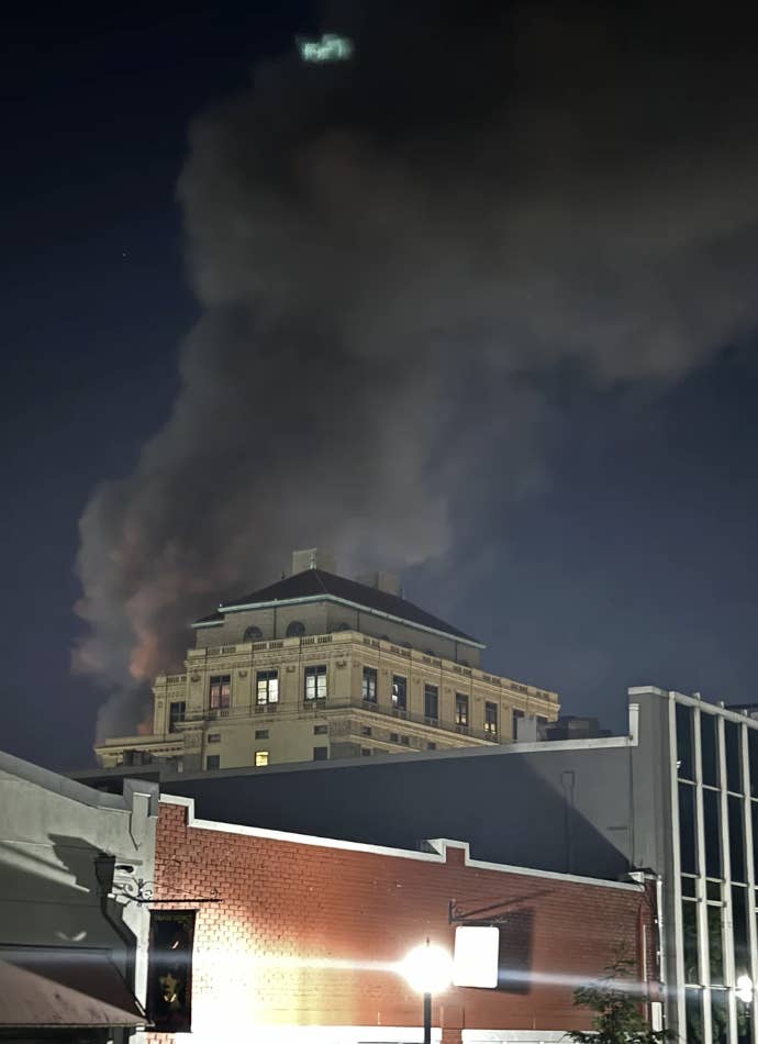 A tall building emits thick, dark smoke from its upper floors at night. Nearby buildings and a streetlight are visible in the foreground