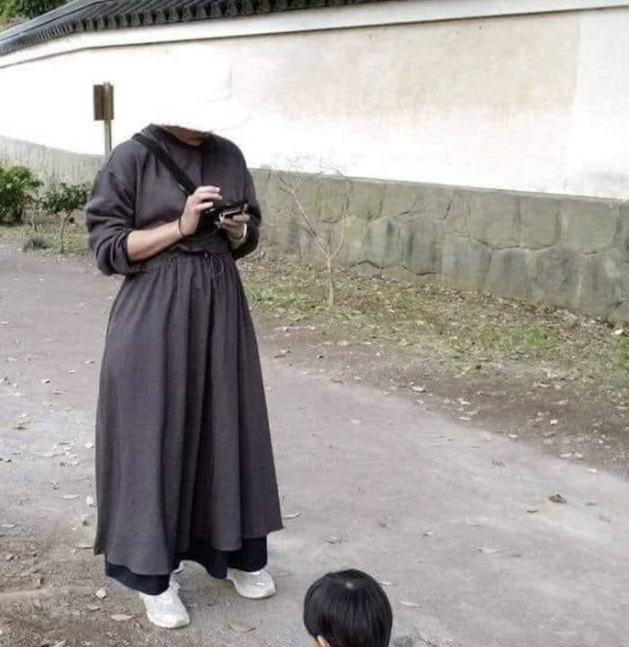 A person in a long dress and hood uses a phone outdoors while a child is playing nearby. The individuals are unidentified