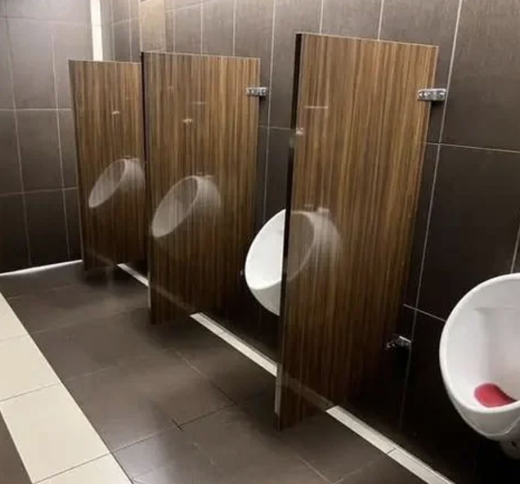 A public restroom with three urinals. The urinal dividers made of wood-like panels partially block the view of each urinal for privacy