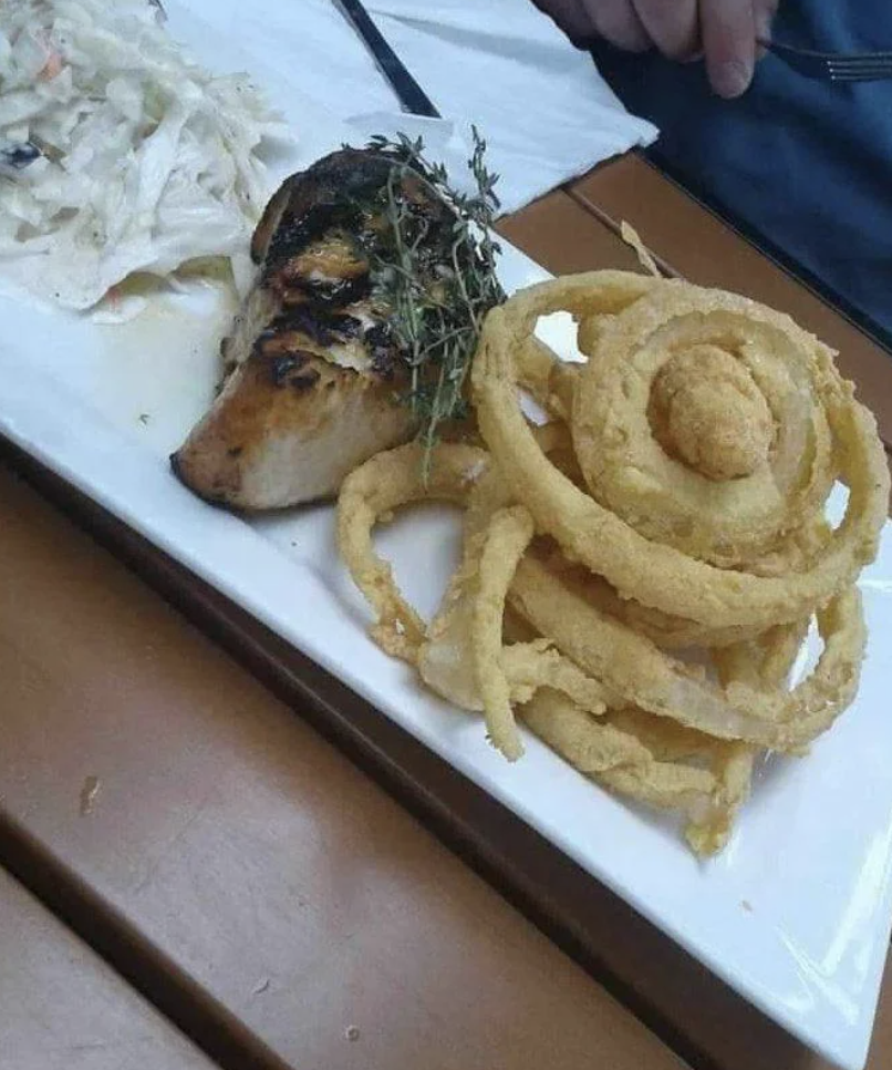 Grilled chicken breast garnished with herbs, a side of onion rings, and a small portion of coleslaw on a rectangular white plate