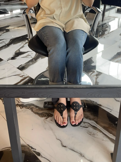 A person in a casual shirt and jeans is sitting on a chair at a salon, with their feet resting on a footrest, wearing sandals with a decorative design