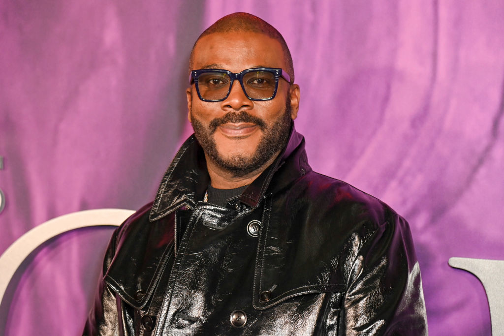 Tyler Perry at an event, wearing a black leather jacket and blue-tinted glasses, smiling at the camera