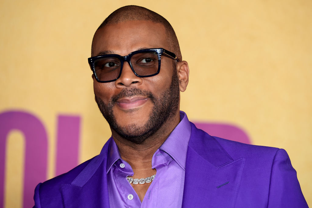 Tyler Perry smiles at an event, wearing a purple suit with a matching shirt, black glasses, and a diamond necklace