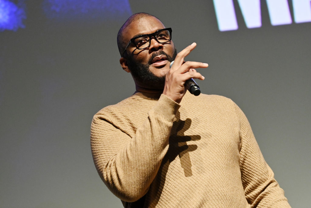 Tyler Perry speaking on stage, holding a microphone, and wearing a textured sweater and glasses