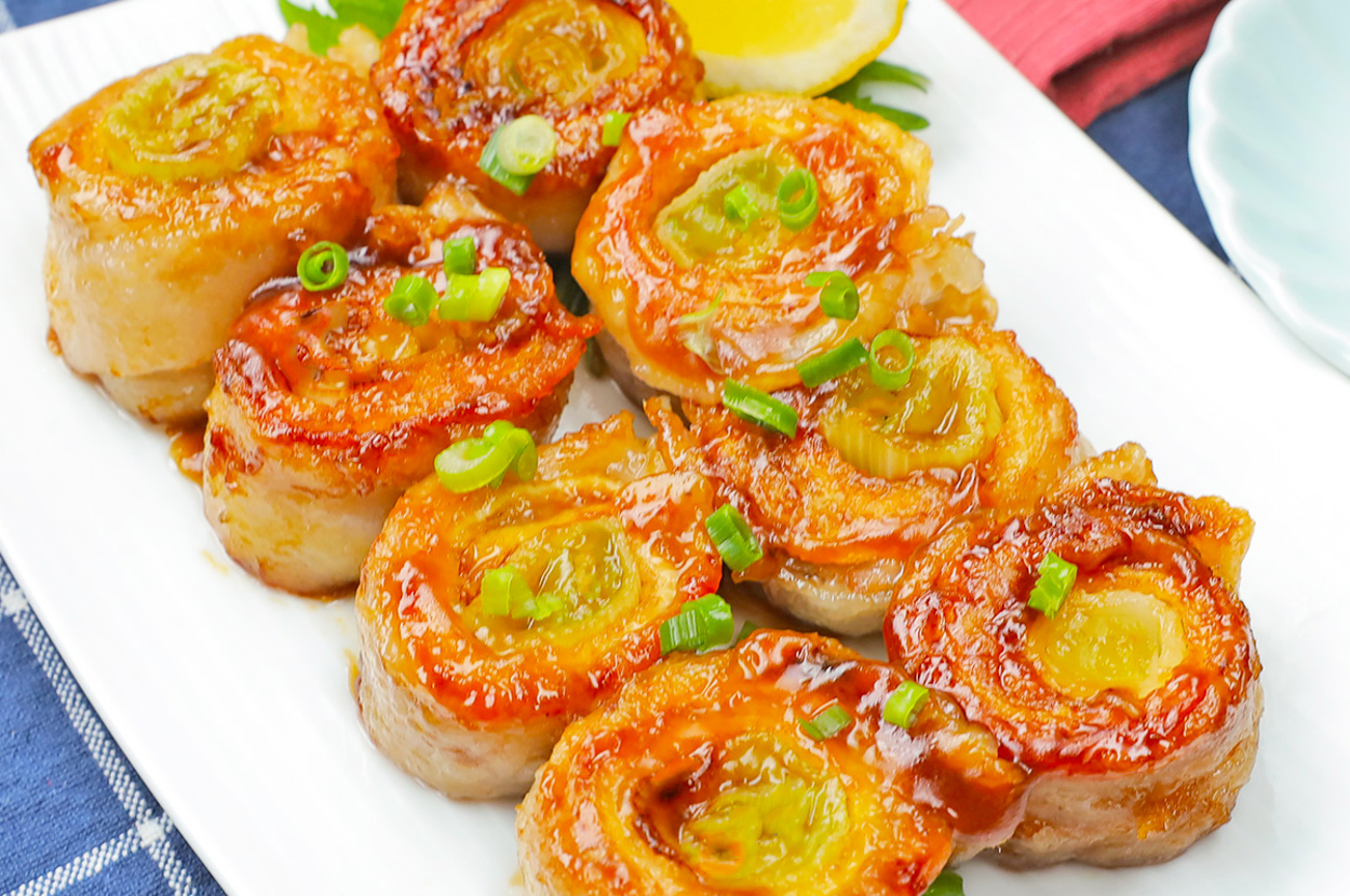 レモンと葉の添え物が付いたお皿に並べられた、タレがかかった豚肉と野菜の巻き物料理。