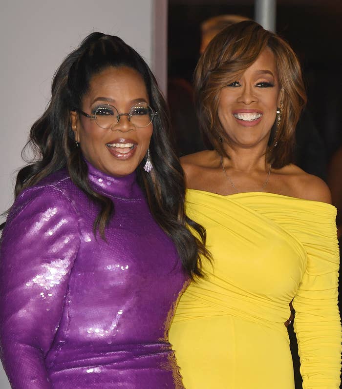 Oprah Winfrey in a sequined dress poses with Gayle King in an off-the-shoulder gown. Gayle is holding a rainbow-colored clutch