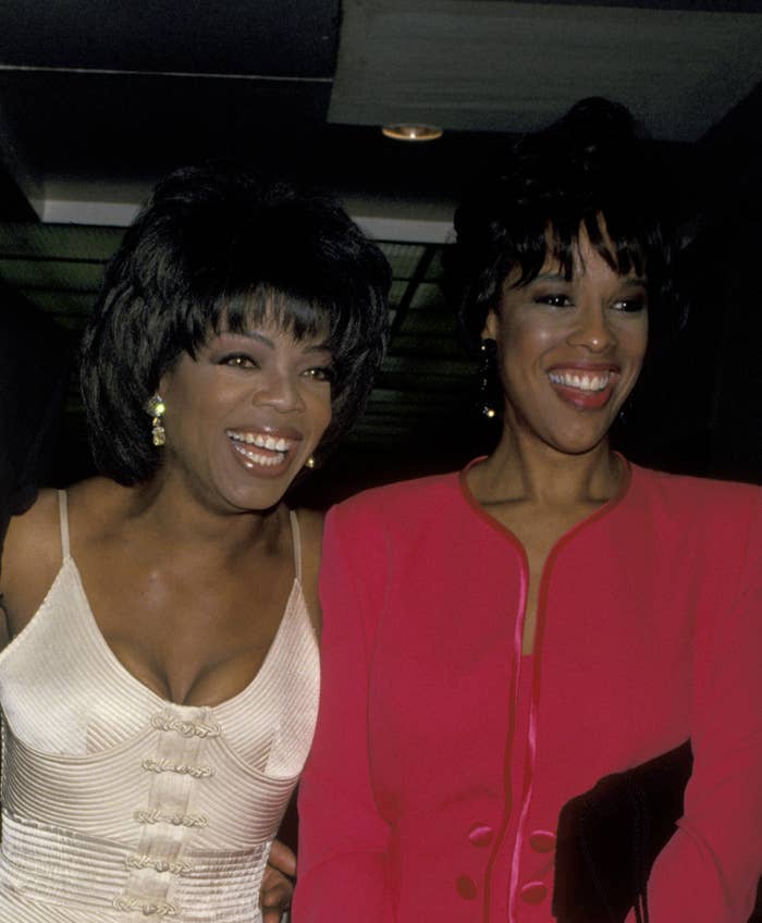 Oprah Winfrey in a floral gown, and Gayle King in a red dress, smiling and standing together