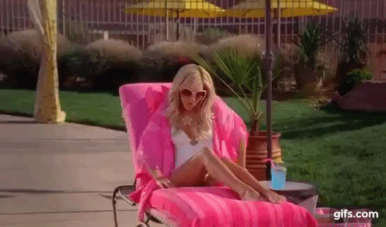 Ashley Tisdale reclines on a sun lounger in a white one-piece swimsuit and large sunglasses, covered with a pink towel. She sips a blue drink by the pool