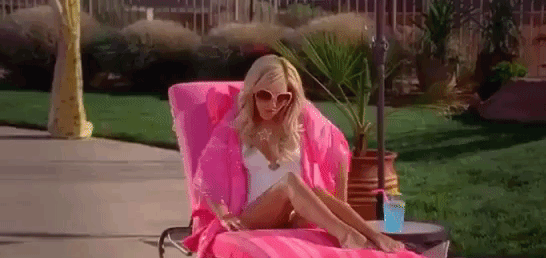 Ashley Tisdale reclines on a sun lounger in a white one-piece swimsuit and large sunglasses, covered with a pink towel. She sips a blue drink by the pool