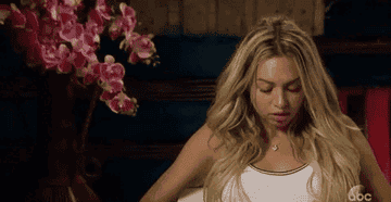 A woman with long blonde hair sits, looking down with a thoughtful expression. There is a vase with pink flowers in the background