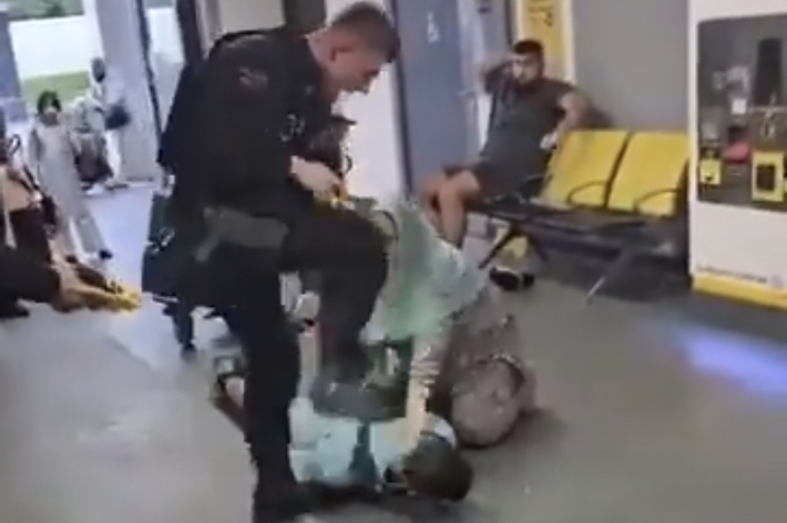 男性の警察官が男性を床に押さえつけている場面が通路で撮影されています。背景に椅子に座っている人々も見られます。