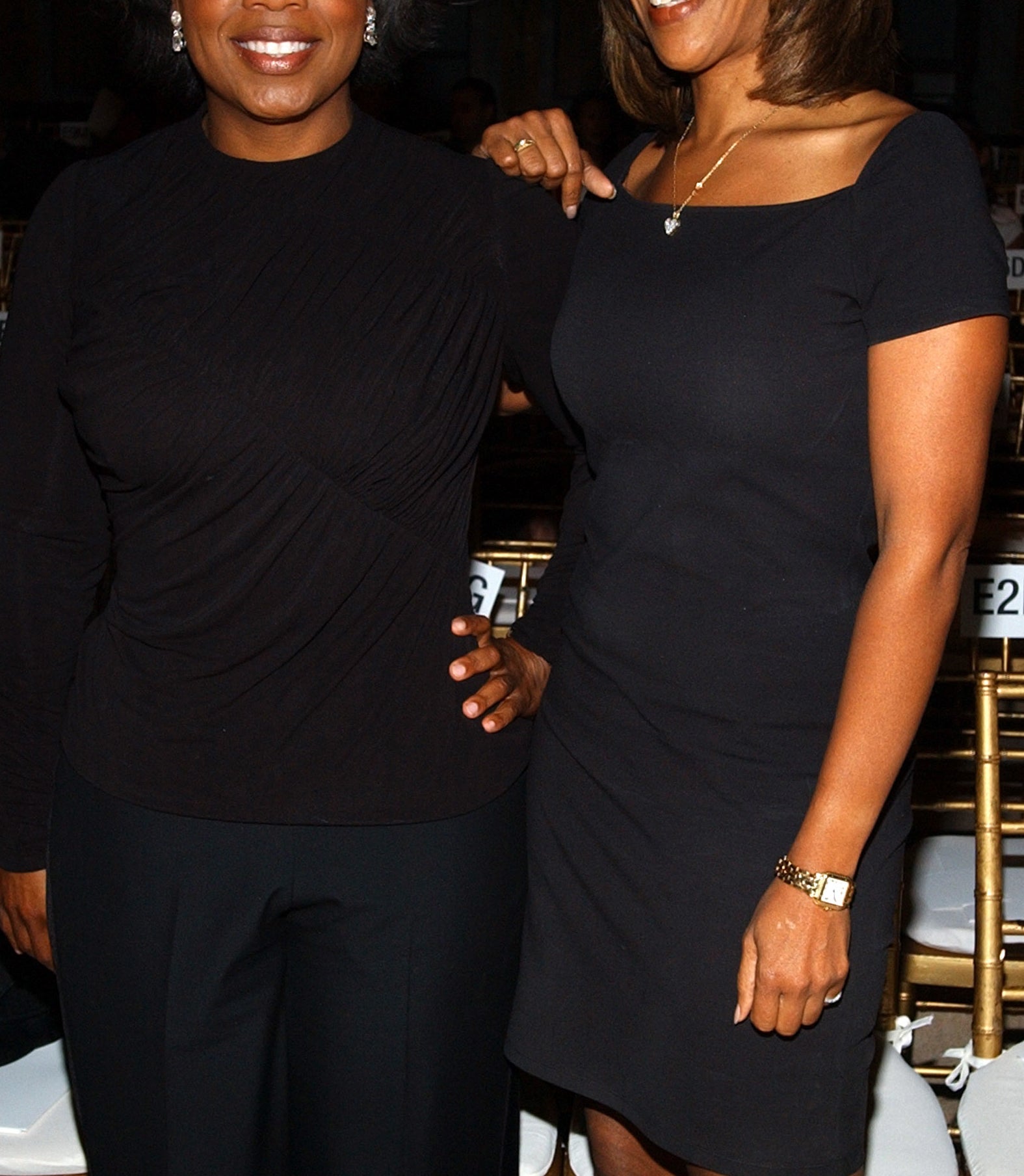 Oprah Winfrey and Gayle King stand together, smiling. Oprah is wearing a long-sleeved black top and pants, while Gayle is dressed in a short-sleeved black dress