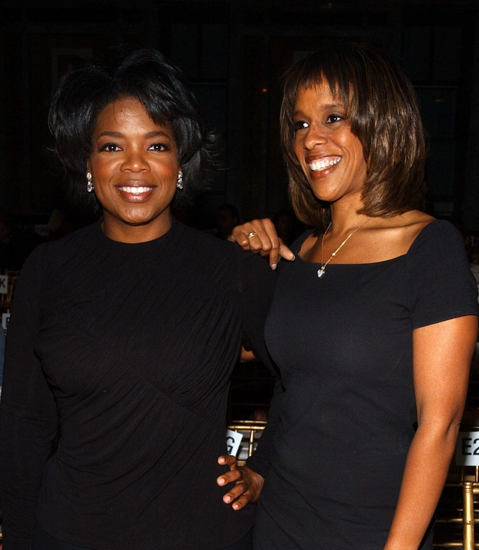 Oprah Winfrey and Gayle King stand together, smiling. Oprah is wearing a long-sleeved black top and pants, while Gayle is dressed in a short-sleeved black dress