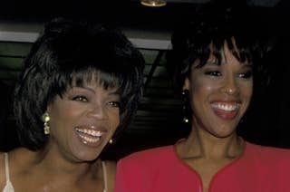 Oprah Winfrey in a floral gown, and Gayle King in a red dress, smiling and standing together