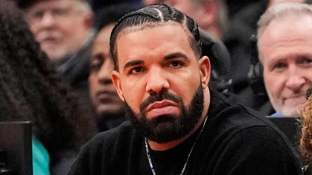 Drake at a public event, wearing a black outfit with a serious expression on his face. Crowd in the background