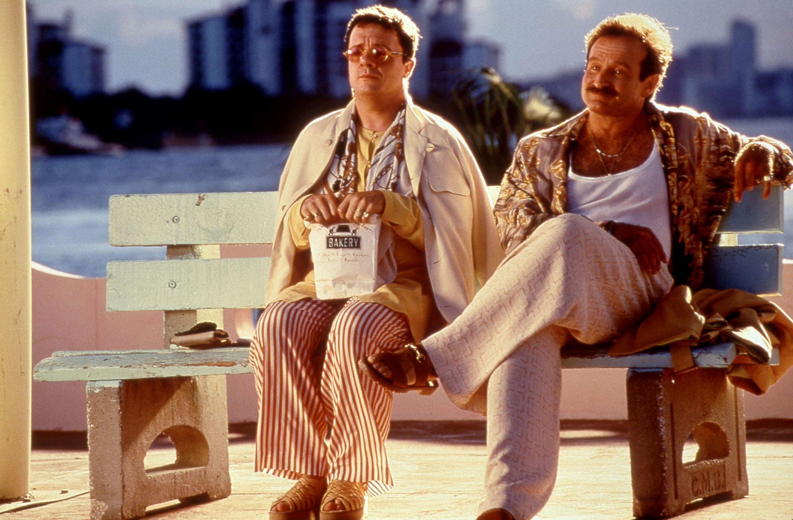 Nathan Lane wearing striped pants and a light jacket sits on a bench with Robin Williams, who is in a tropical shirt and light trousers in a scene from "The Birdcage."