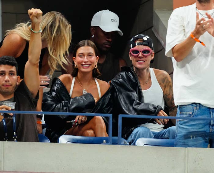 Hailey Bieber smiles and sits next to Justin Bieber, who wears a leather jacket, tank top, and pink sunglasses, at a sports event