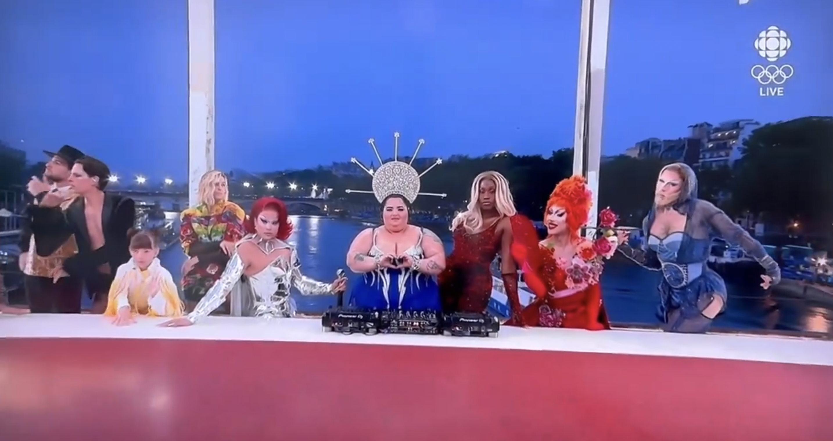 Drag queens in extravagant outfits pose with a child and a man in a tuxedo at a DJ booth, during a live televised event