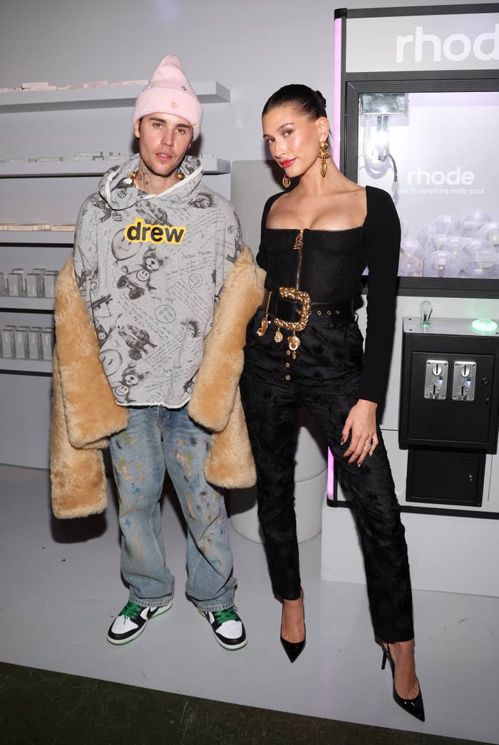 Justin Bieber in a casual outfit with a graphic hoodie and beanie, and Hailey Bieber in a chic dark ensemble with stylish accessories at a Rhone event