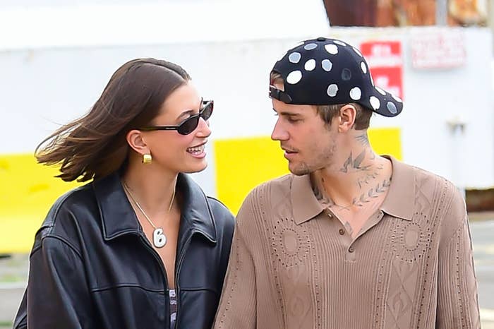 Hailey Bieber, wearing a black leather jacket, smiles at Justin Bieber, who has a polka-dot cap and a beige knit sweater