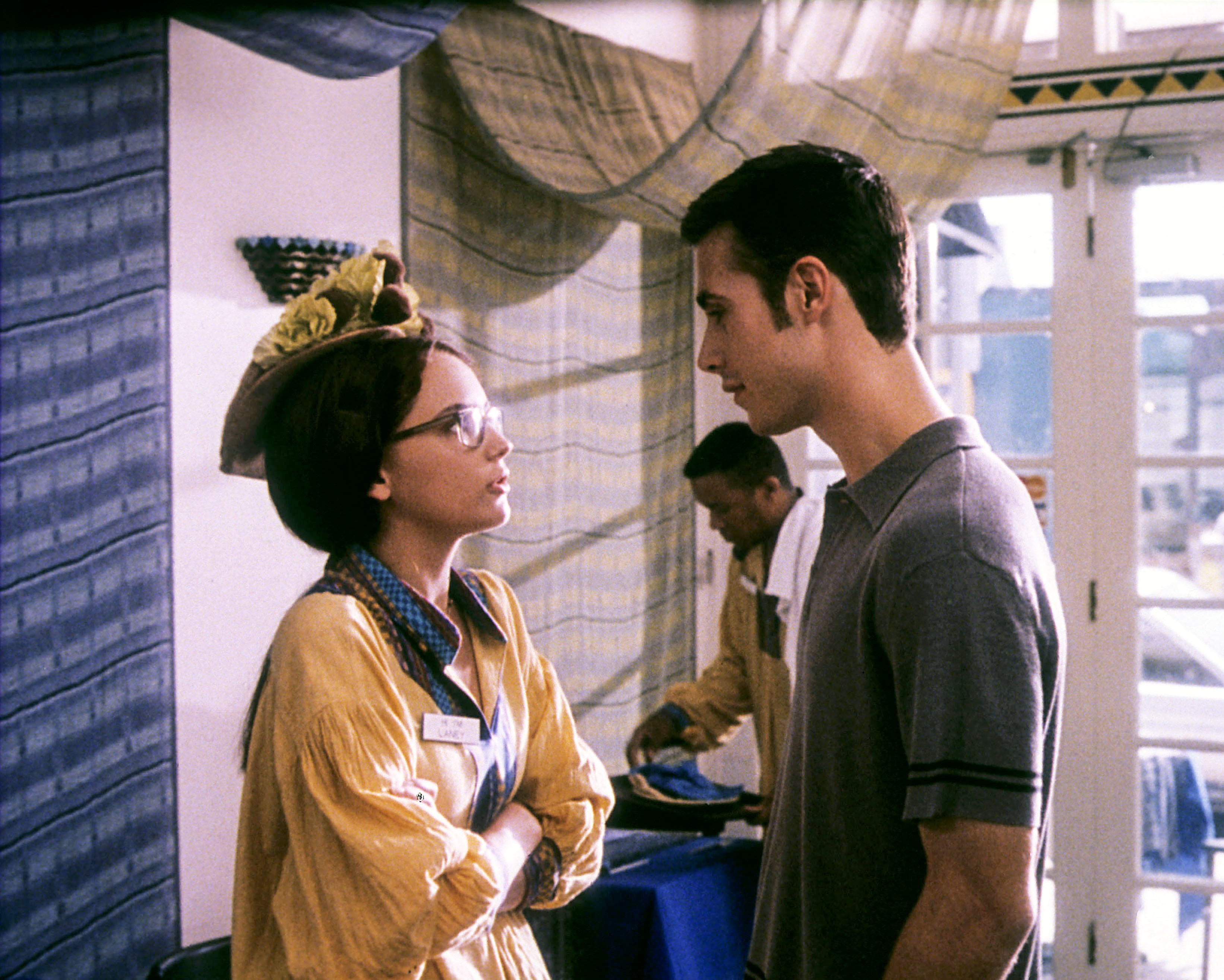 Rachael Leigh Cook and Freddie Prinze Jr. are having a conversation in a room decorated with patterned curtains. Both wear casual outfits