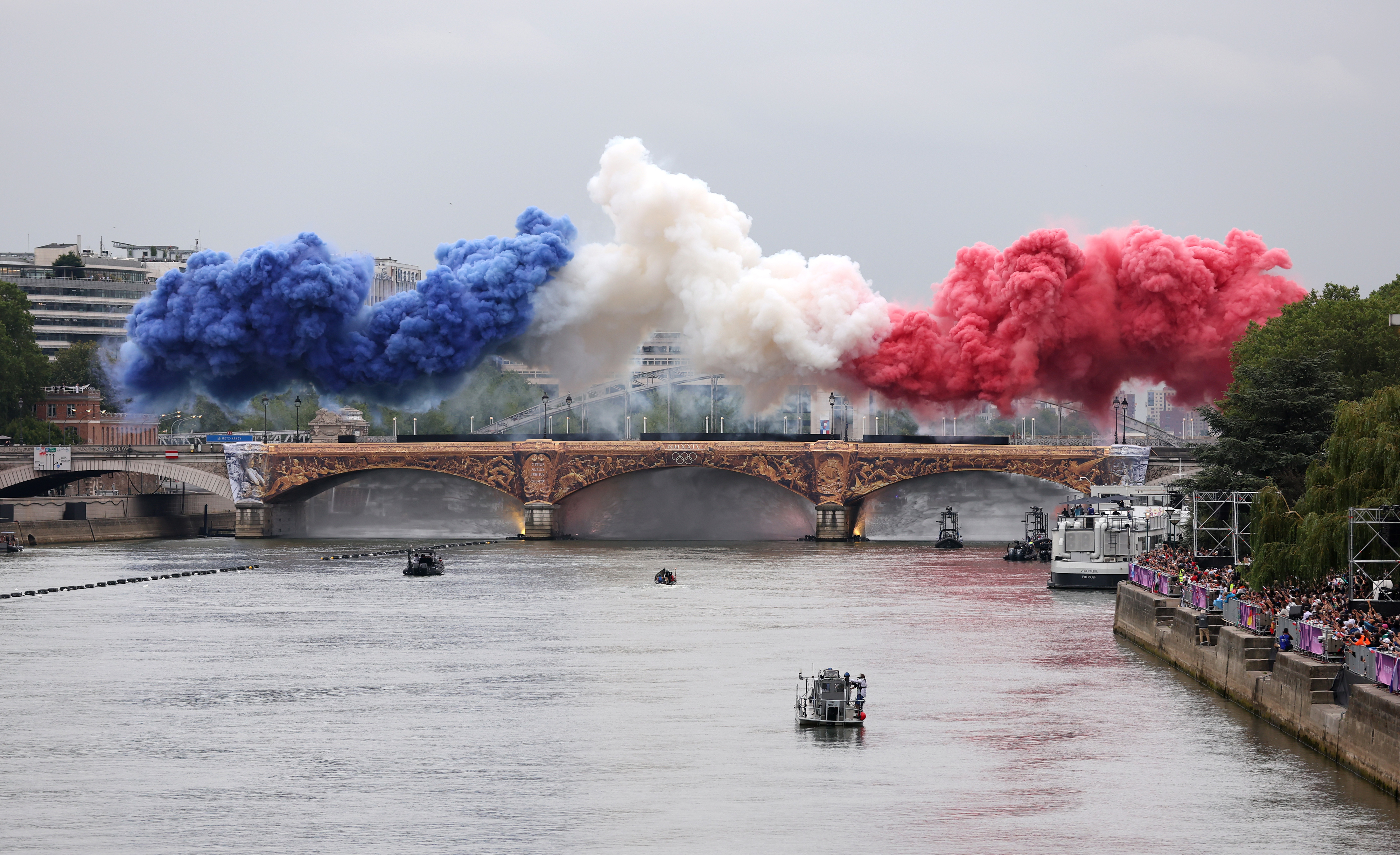 Paris bridge with smoke in blue, white, and red colors overhead. Crowds watch on both riverbanks. No people in close-up view