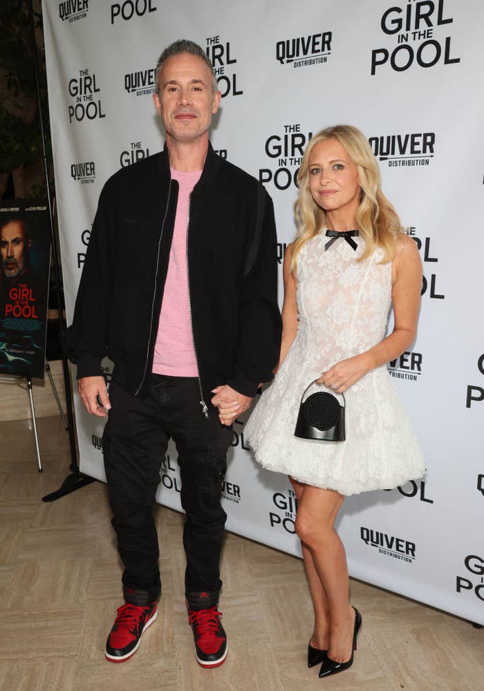 Freddie Prinze Jr. and Sarah Michelle Gellar holding hands at a "The Girl in the Pool" event. The man is dressed casually, and the woman is wearing a short lace dress