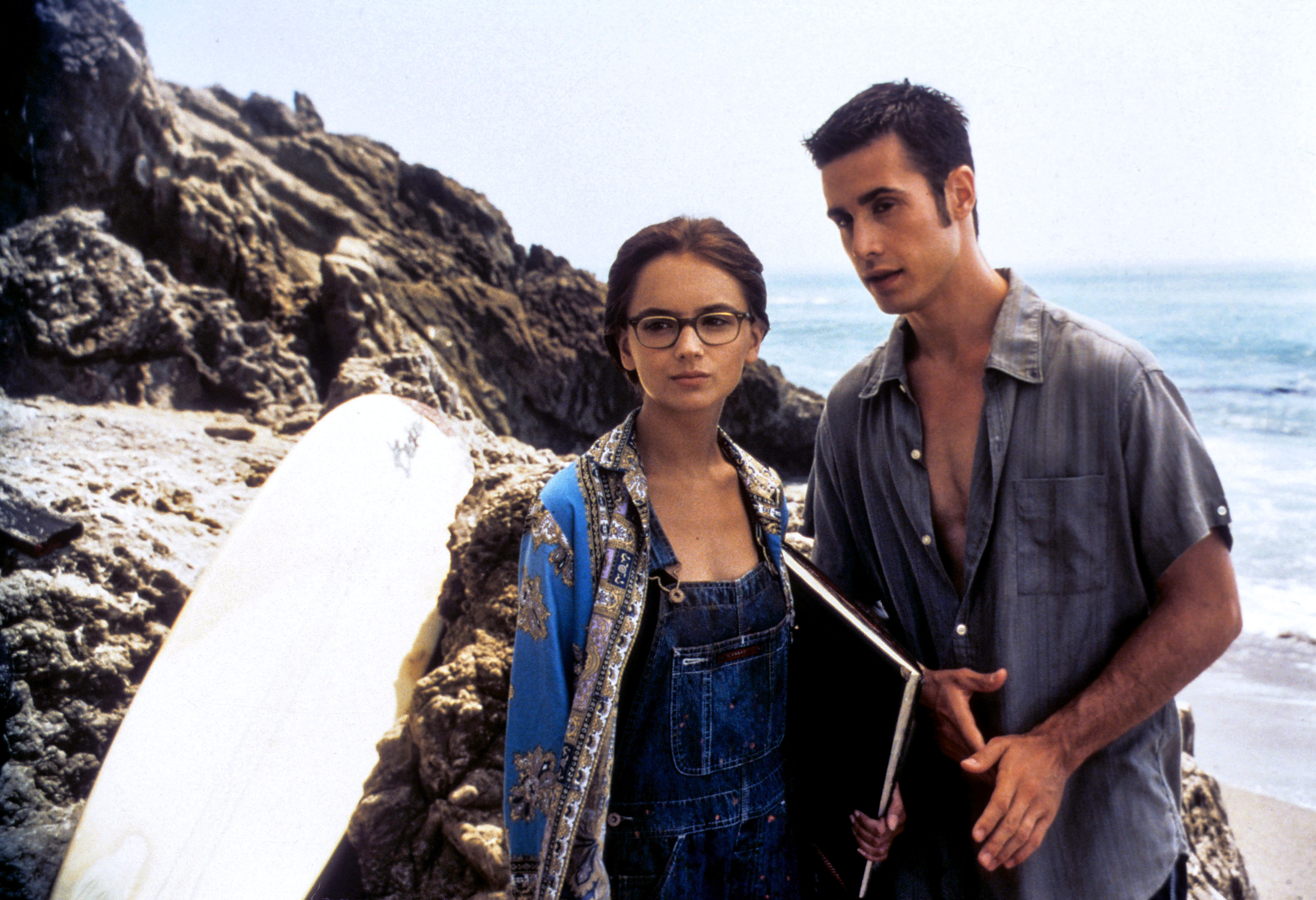 Rachel Leigh Cook in glasses and overalls with Freddie Prinze Jr. wearing a casual shirt, standing near a surfboard and rocky shore in a beach scene from a movie