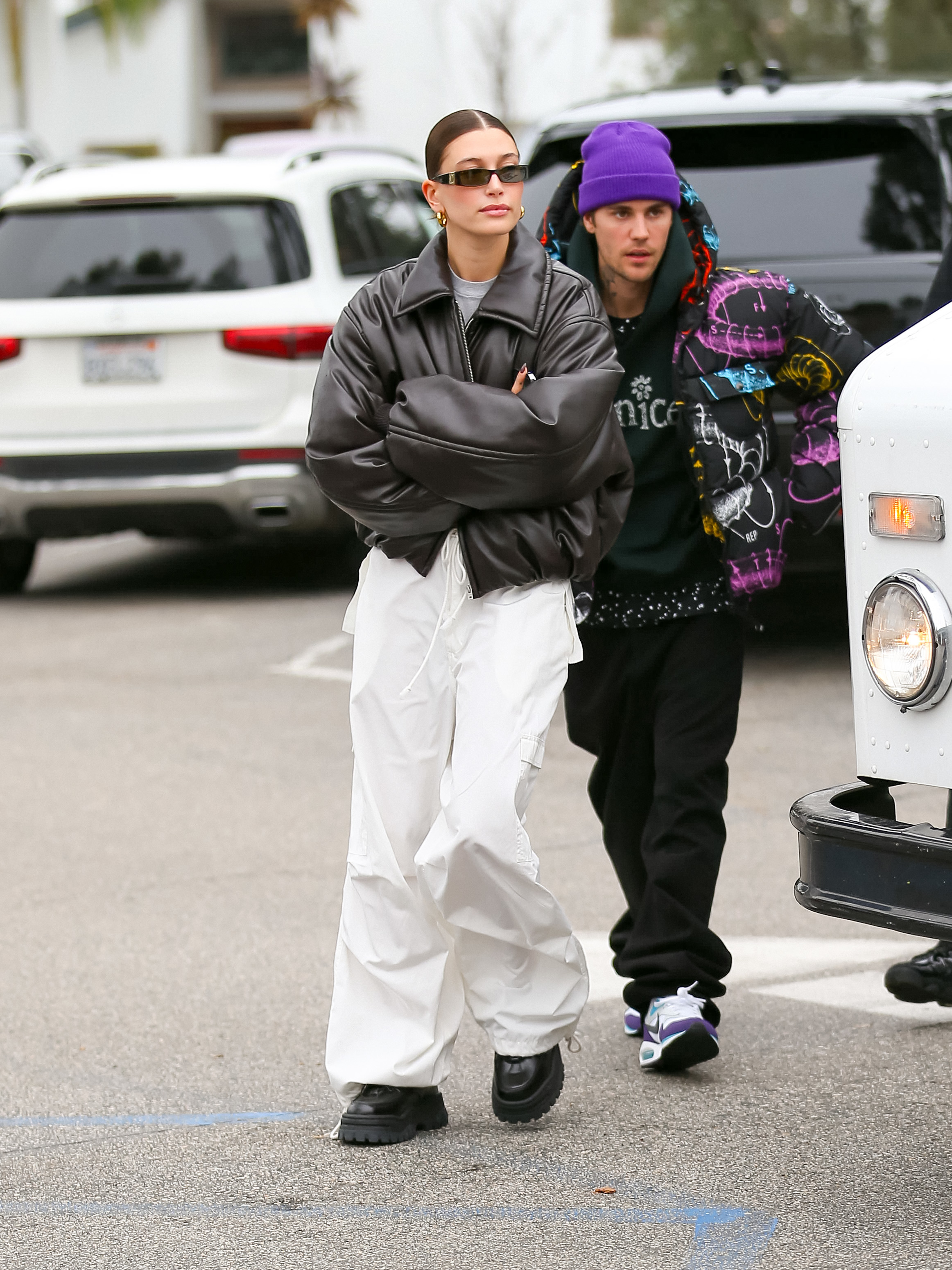 Hailey Bieber and Justin Bieber are seen walking. Hailey wears oversized white pants and a leather jacket, while Justin wears a colorful hoodie and a purple beanie