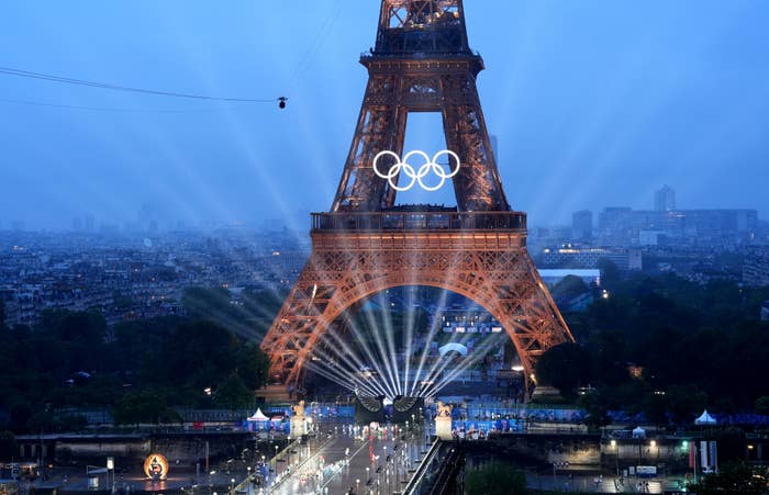 The Eiffel Tower illuminated with Olympic rings as beams of light shine down. The scene captures an event set up at the base with a cityscape in the background