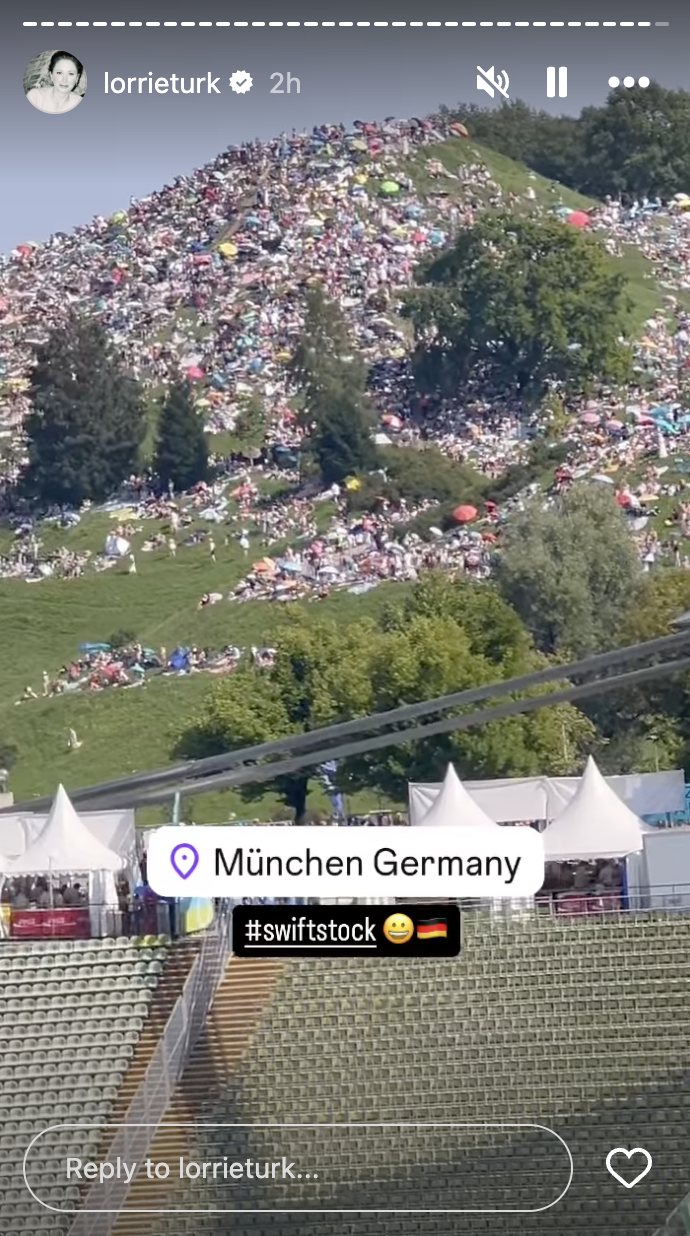 People gathering on a large, grassy hill in München, Germany, for an event labeled #swiftstock. Tents and trees are visible, and an empty seating area is in the foreground