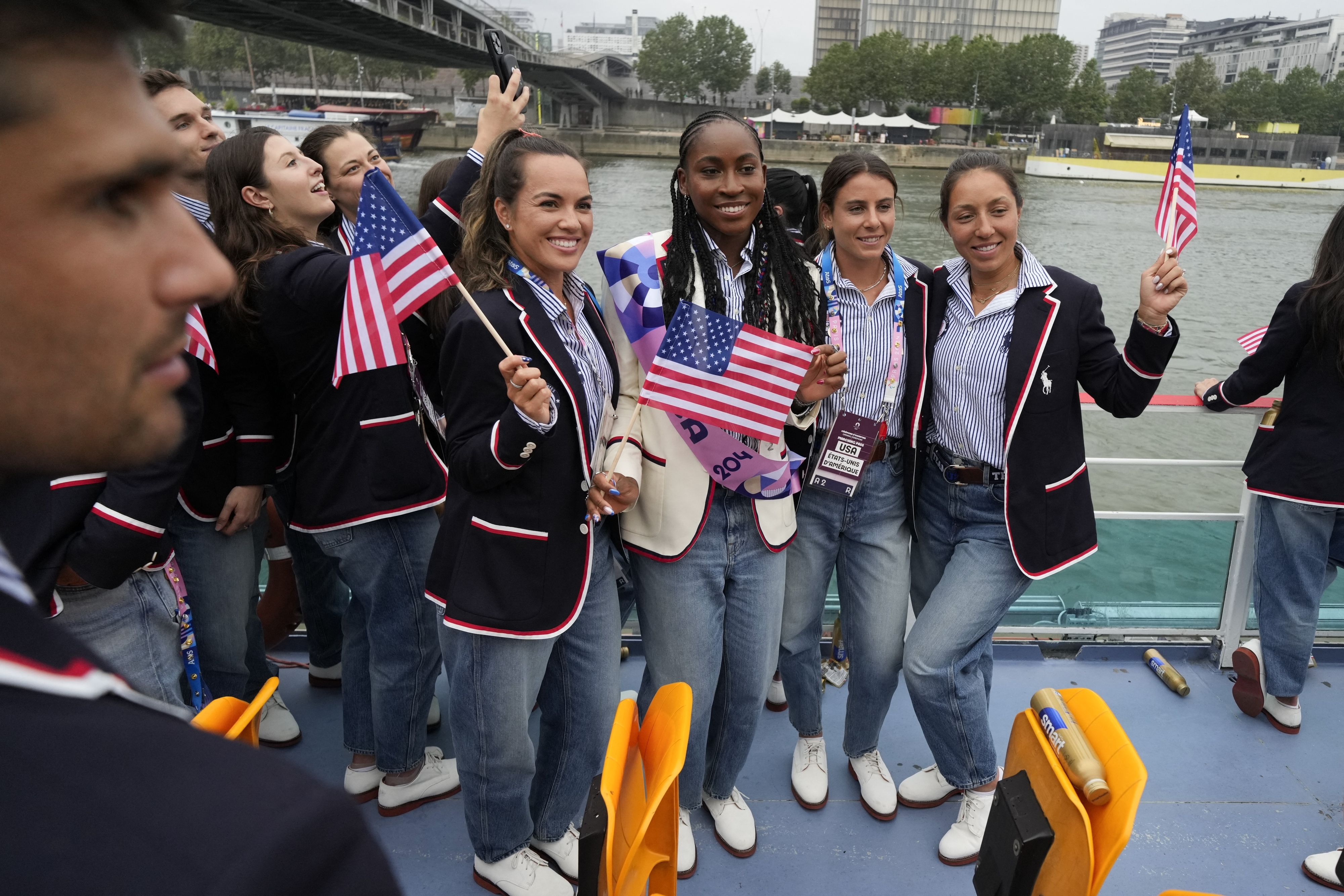 Group of athletes, including Coco Gauff, wearing USA blazers with American flags on a boat, celebrating in a city with buildings and a bridge in the background
