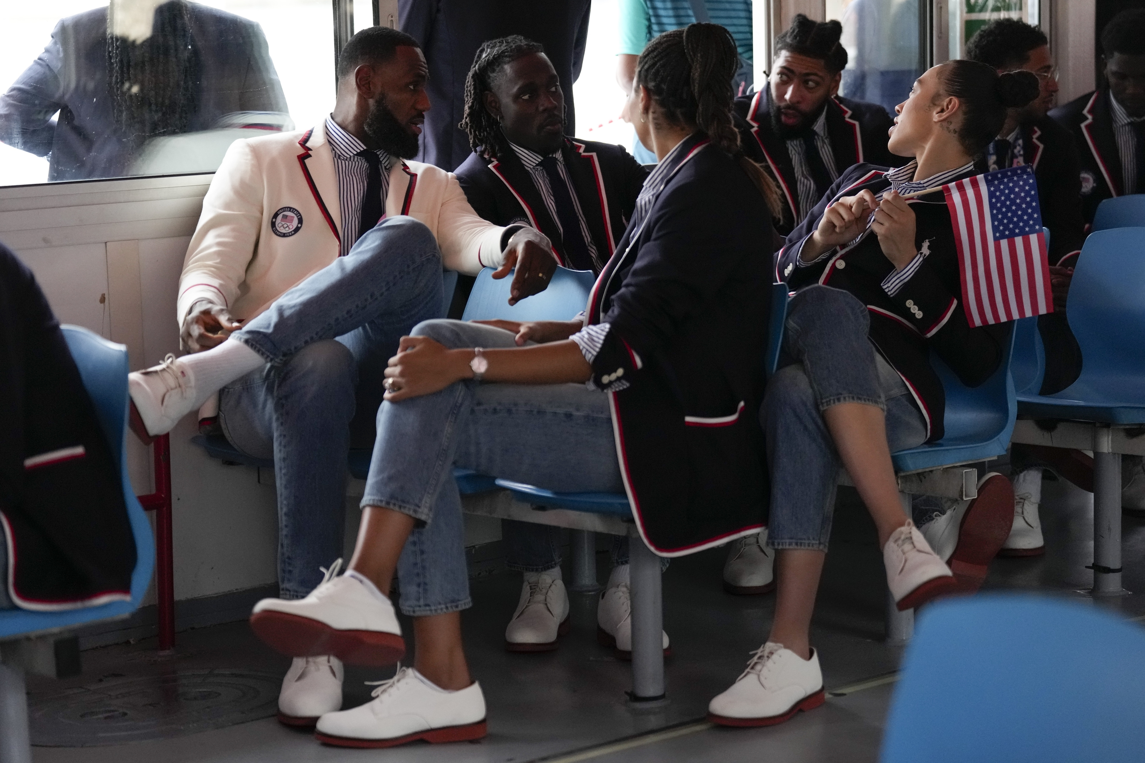 Several individuals sit and talk, wearing matching casual outfits with white blazers, blue jeans, and white shoes. One person holds an American flag